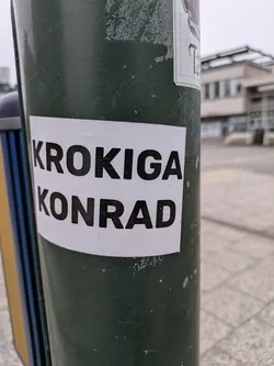 A rectangular sticker with a white background and black text reading 'KROKIGA KONRAD' is adhered to a green metal pole outdoors. The sticker appears to be in good condition. There are other stickers on the same pole, though the text is not visible. The background includes a paved area and a building.