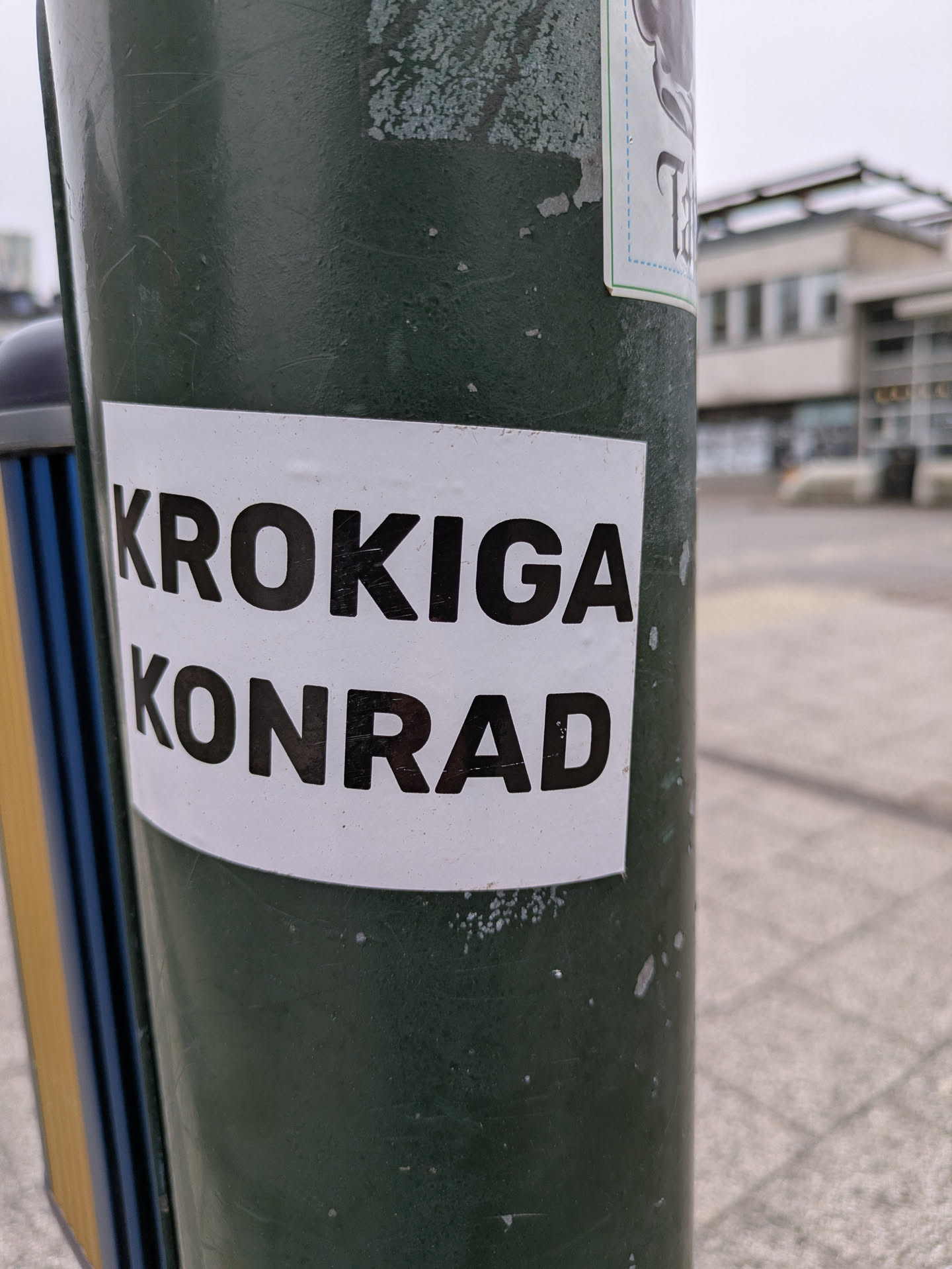 A rectangular sticker with a white background and black text reading 'KROKIGA KONRAD' is adhered to a green metal pole outdoors. The sticker appears to be in good condition. There are other stickers on the same pole, though the text is not visible. The background includes a paved area and a building.