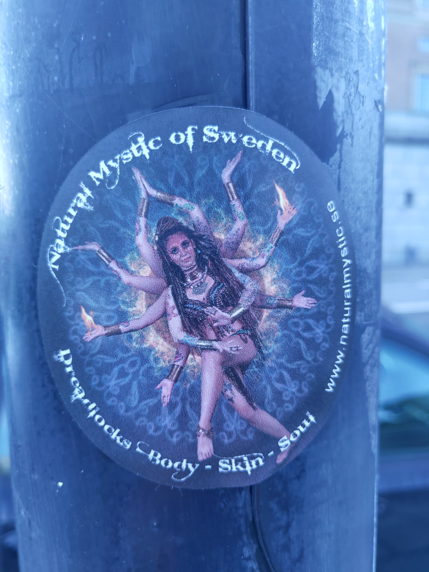 Round sticker featuring an illustration of a woman with multiple arms, reminiscent of a Hindu goddess, in a cosmic mandala. Text on the sticker includes the brand name, location, and a tagline emphasizing dreadlocks, body, skin, and soul.