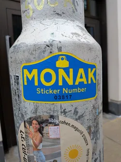 The central sticker is a die-cut design with a prominent blue background and a bright yellow border. At the top center, there is a yellow icon resembling a camera or a security device. Below this icon, the word 'MONAK' is displayed in large, bold yellow capital letters. Further below, in smaller yellow text, it reads 'Sticker Number', followed by the black numerical identifier '03817'. The sticker is applied to a textured, weathered grey pole.