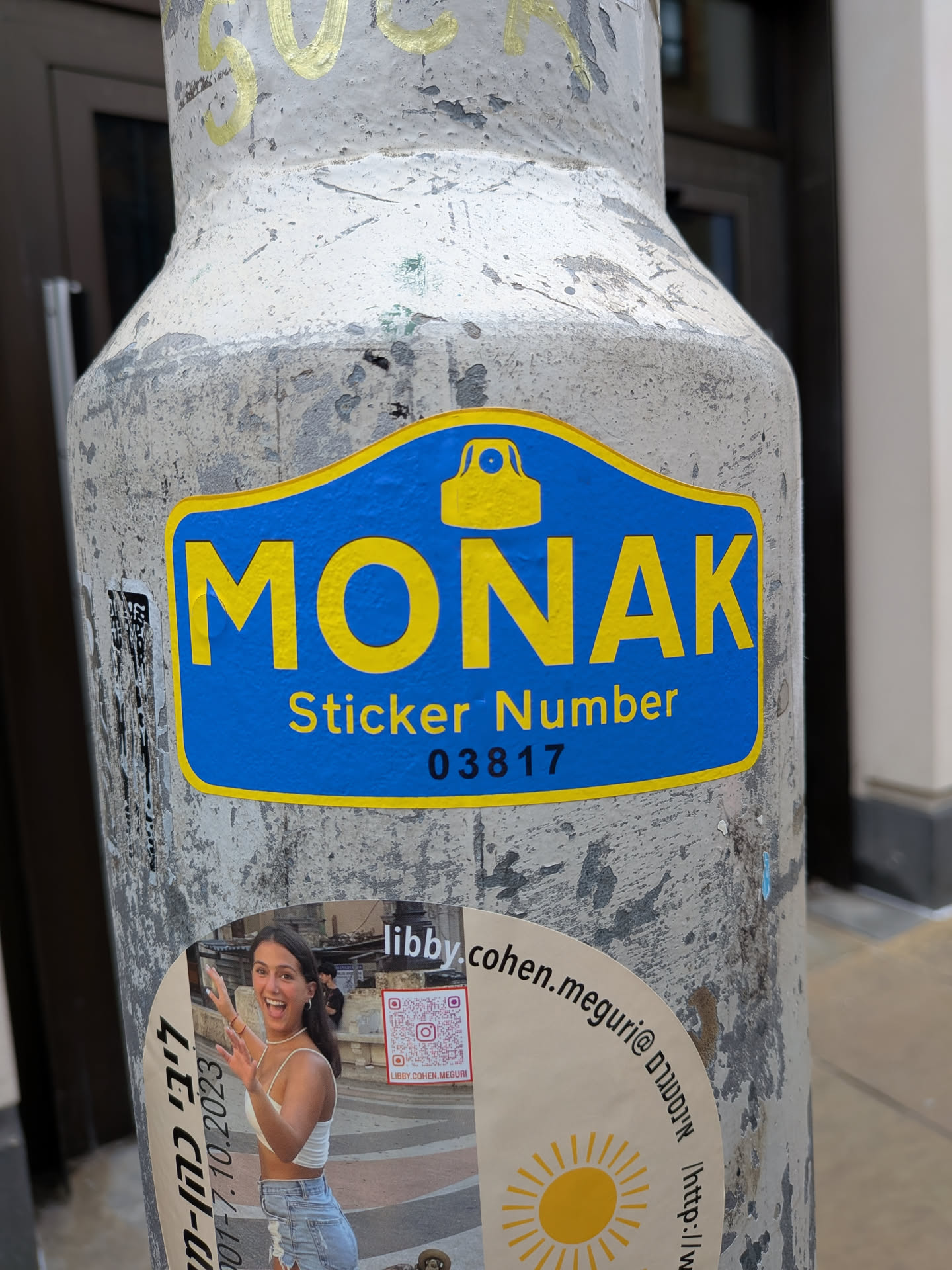The central sticker is a die-cut design with a prominent blue background and a bright yellow border. At the top center, there is a yellow icon resembling a camera or a security device. Below this icon, the word 'MONAK' is displayed in large, bold yellow capital letters. Further below, in smaller yellow text, it reads 'Sticker Number', followed by the black numerical identifier '03817'. The sticker is applied to a textured, weathered grey pole.