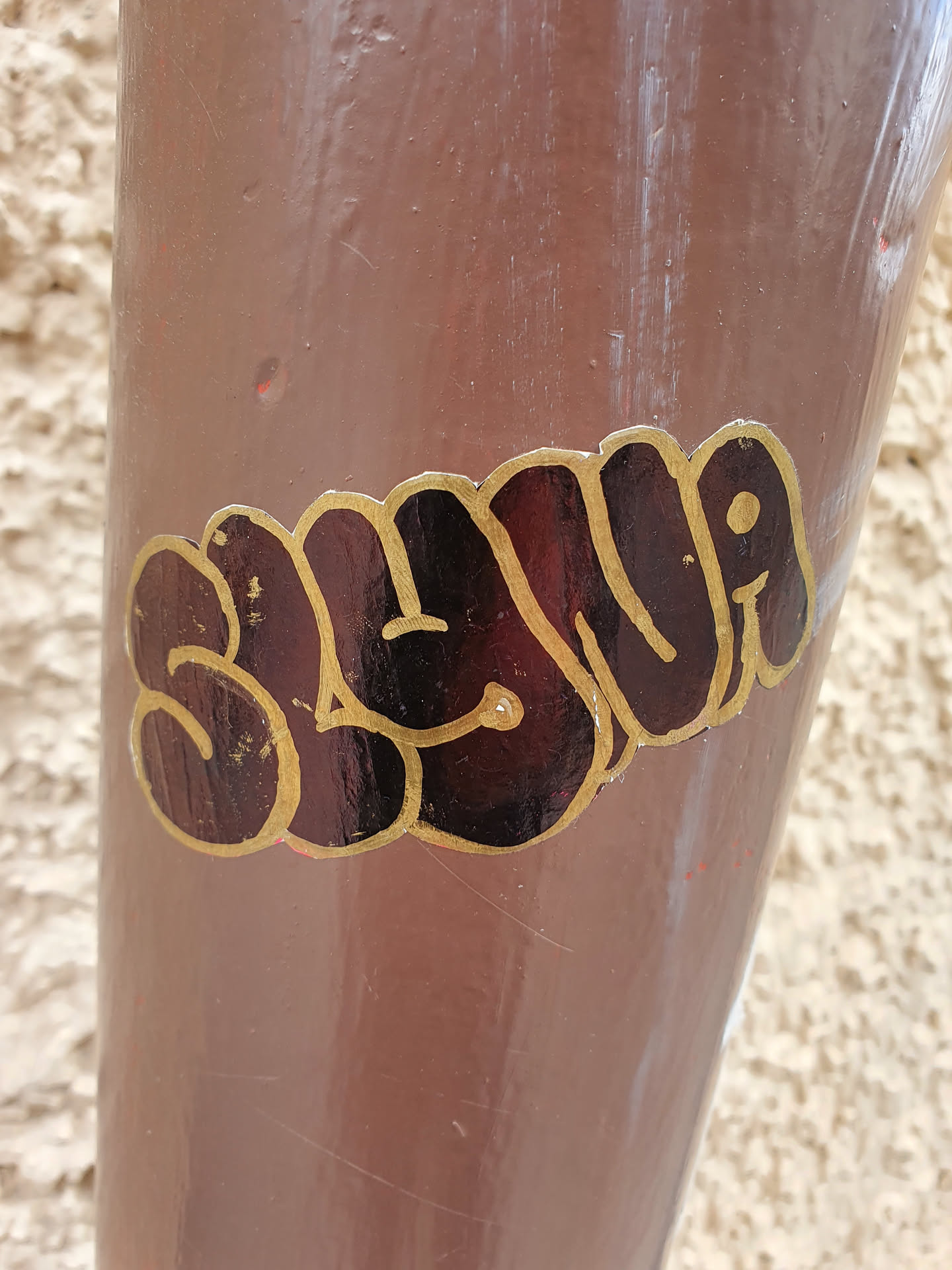A close-up shot of a black and gold sticker featuring the name "Slyvia" in a stylized bubble-letter font. The sticker is affixed to a brown cylindrical surface, possibly a pole or pipe, outdoors. The letters have a glossy finish, and the gold trim adds a touch of elegance. The overall style of the sticker is reminiscent of graffiti art or street art.