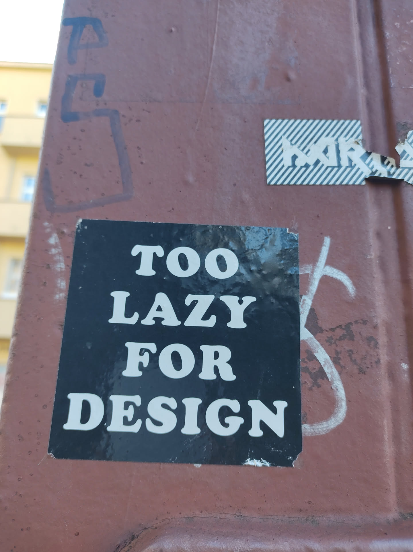 A black sticker with white text on a brown surface. The text says "TOO LAZY FOR DESIGN".