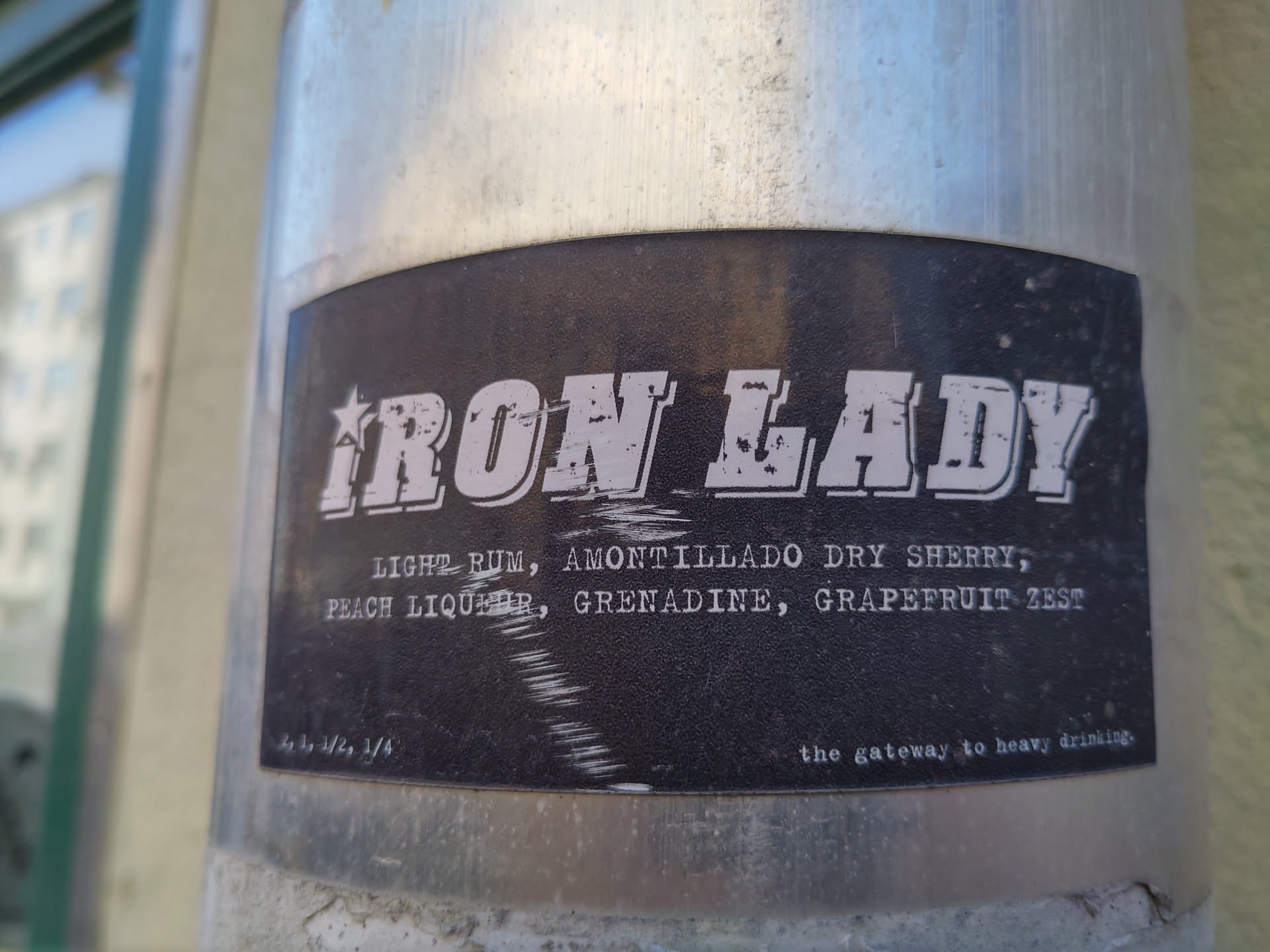 A black sticker with white text.  It says "IRON LADY" in a large, stylized font. Below that, a list of ingredients: Light Rum, Amontillado Dry Sherry, Peach Liqueur, Grenadine, Grapefruit Zest. At the bottom it says, "the gateway to heavy drinking".