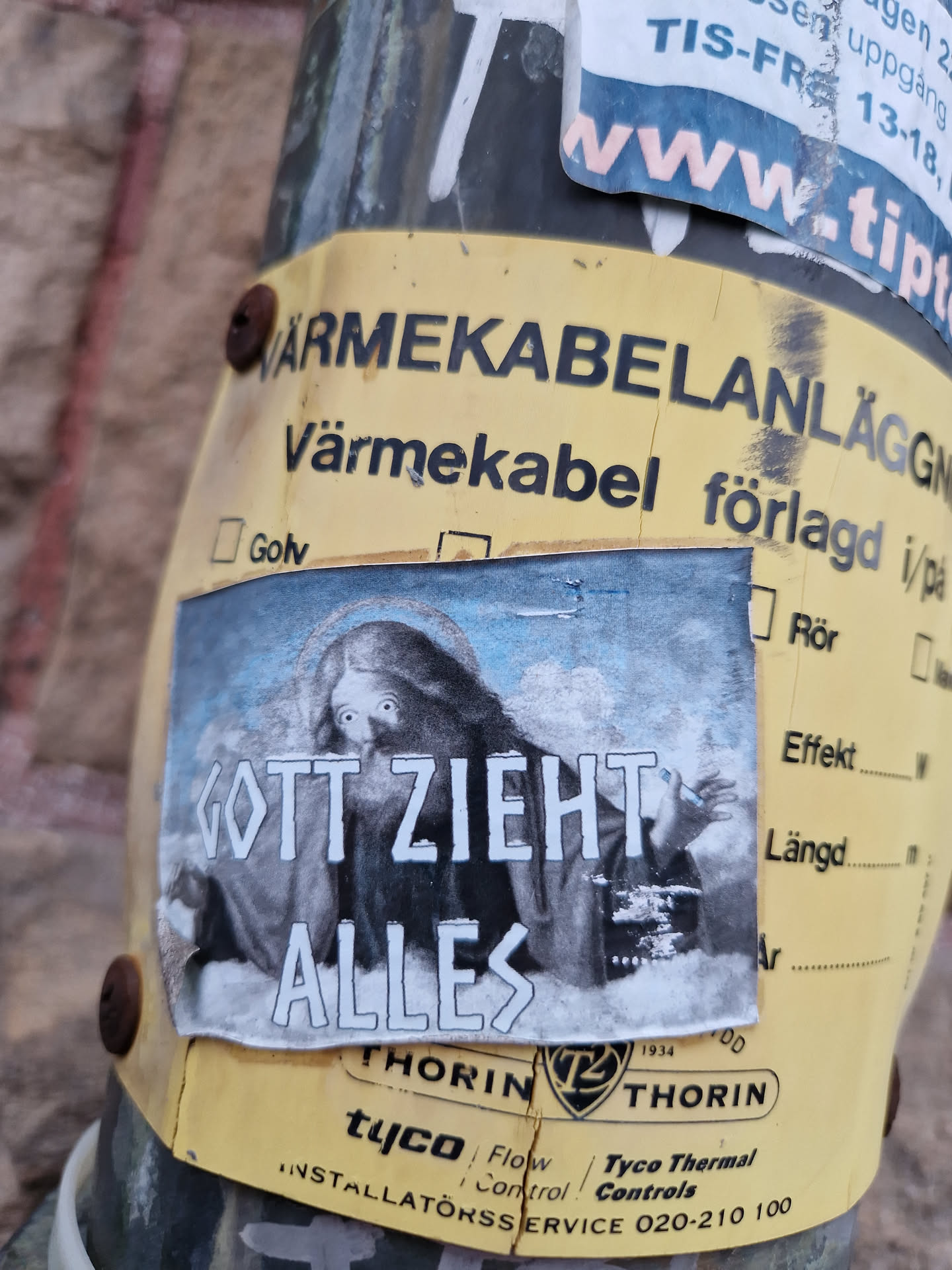 A sticker featuring a grayscale image of a hooded figure with the German phrase "Gott zieht alles" (God pulls everything) overlaid. The sticker is affixed to a pole with other labels.