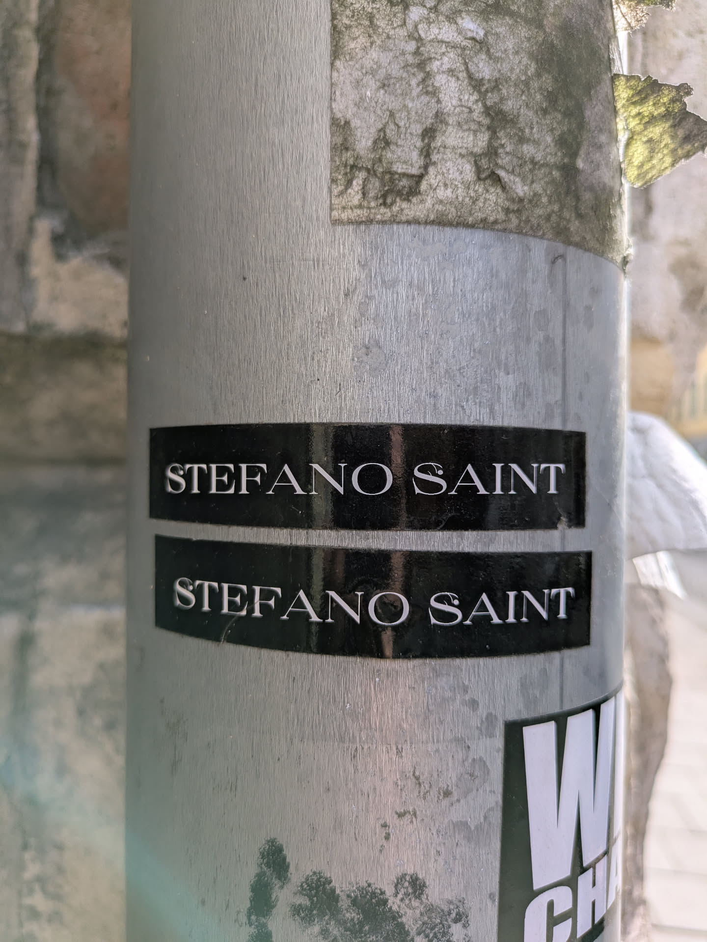 The image features a sticker adhered to a metallic pole. The sticker has a black background with the text "STEFANO SAINT" printed in white. There are two identical stickers placed one above the other. Below those there is another sticker that partially appears in the frame