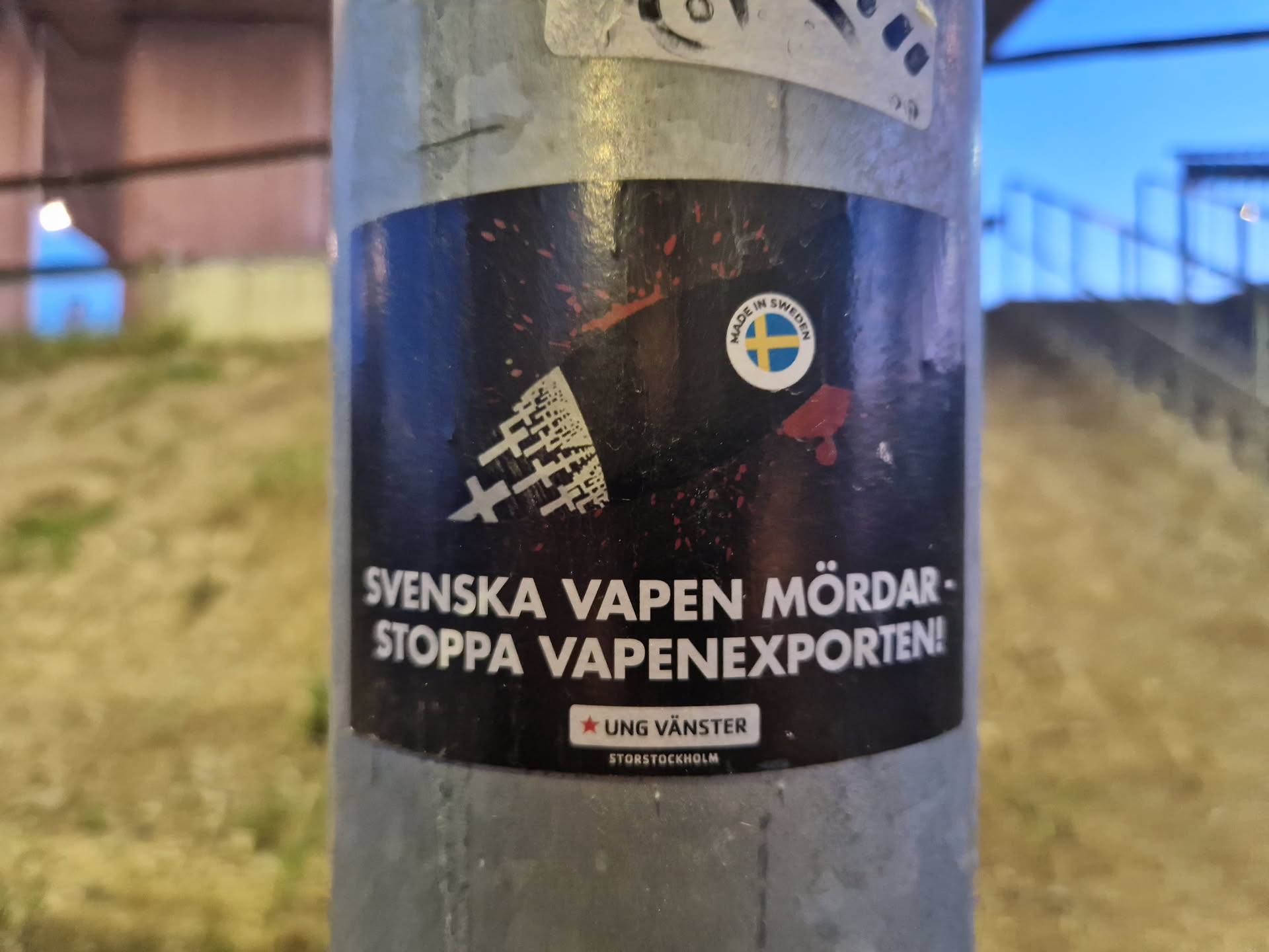 A sticker with a dark background showing a silhouette of crosses and a blood drop. It has text in Swedish that translates to "Swedish weapons kill - Stop the arms export!"  There's a small Swedish flag and the logo of Ung Vänster (Young Left) Stockholm.
