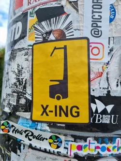 Yellow sticker with a black pictogram of what seems to be a type of vehicle and the text X-ING. The sticker is attached to a pole covered with other stickers.