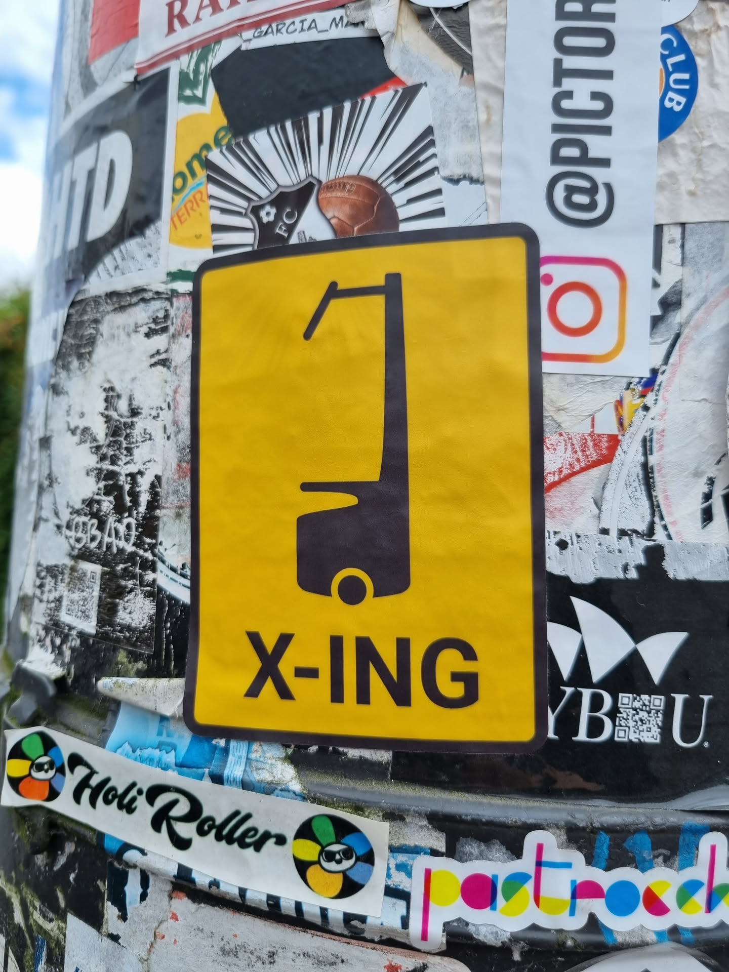 Yellow sticker with a black pictogram of what seems to be a type of vehicle and the text X-ING. The sticker is attached to a pole covered with other stickers.