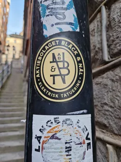 A sticker featuring a logo for Aktiebolaget Black & Navy, which seems to be an electric tattooing company. The logo is a circular design with the company name written around it and stylized initials in the center. The sticker is black and yellow. 