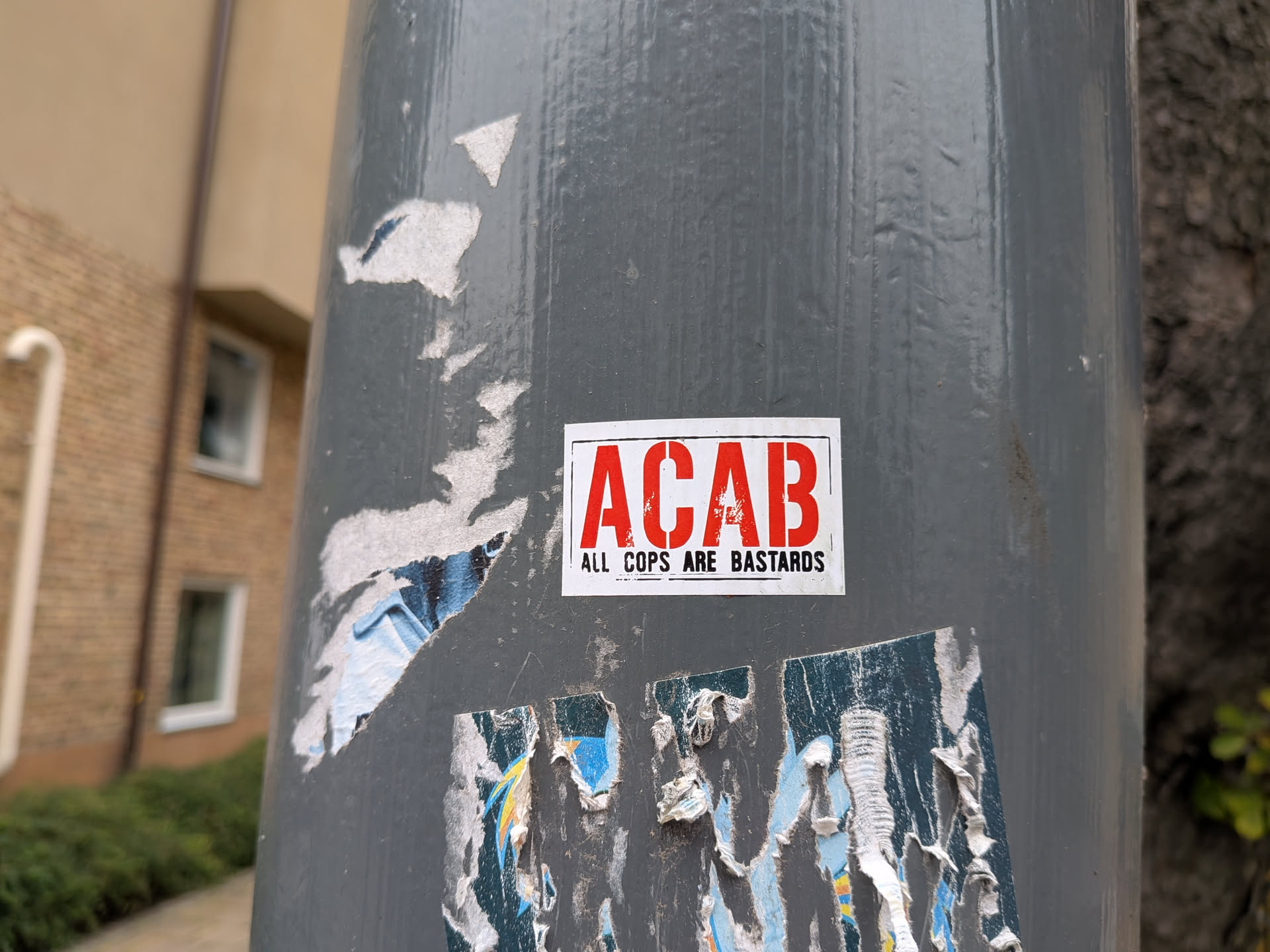 The sticker is rectangular with a white background and a red border. Prominently displayed in large, red, sans-serif capital letters are the letters 'ACAB'. Below this, in smaller black sans-serif capital letters, is the text 'ALL COPS ARE BASTARDS'. The sticker appears to be slightly worn at the edges and is affixed to a grey, cylindrical pole, with other torn and weathered stickers surrounding it.