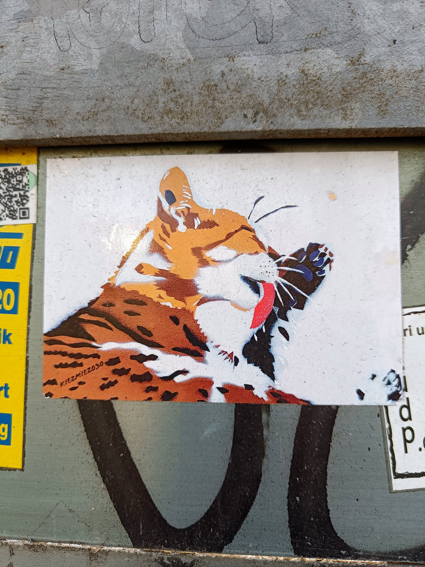 A sticker featuring a stylized illustration of an orange tabby cat cleaning its paw. The cat's fur is depicted with a mix of orange, white, and brown tones, using a stencil-like technique. The artist's signature, "KIRZMIT2030," is visible in the bottom left corner. The sticker has a slightly distressed, street art-like aesthetic.