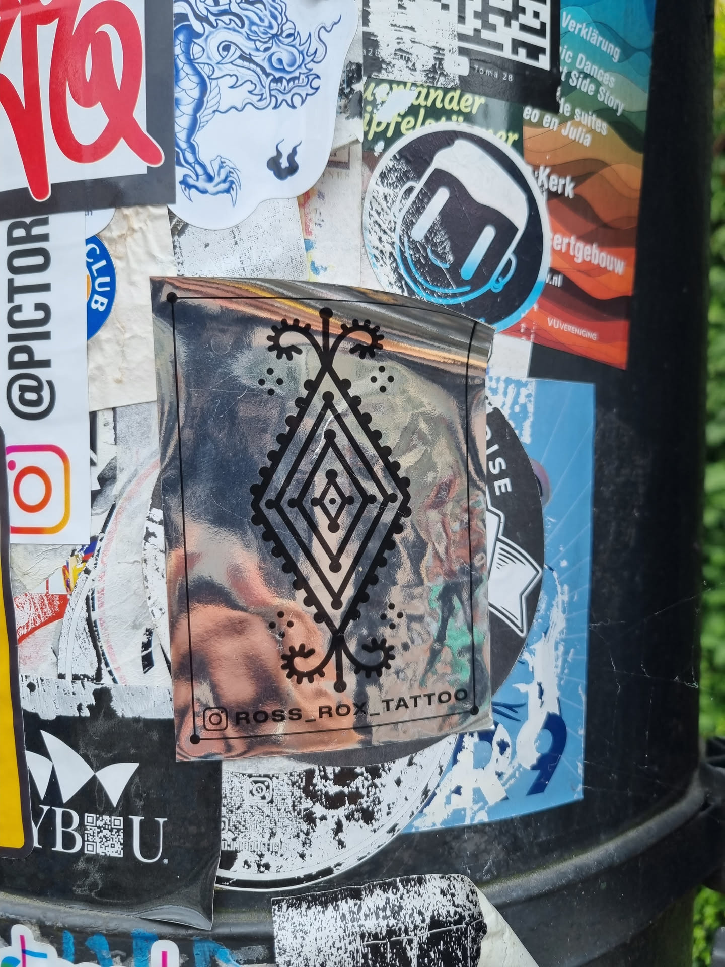 A holographic silver sticker with a black geometric design. The sticker features a central diamond shape with ornate detailing and smaller decorative elements.  The text "ROSS_ROX_TATTOO" is visible at the bottom, indicating the artist or studio.