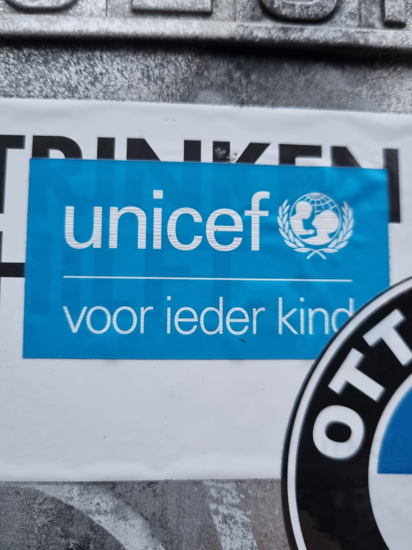 A blue UNICEF sticker with the UNICEF logo and the text "voor ieder kind" (Dutch for "for every child") is affixed to a surface. 