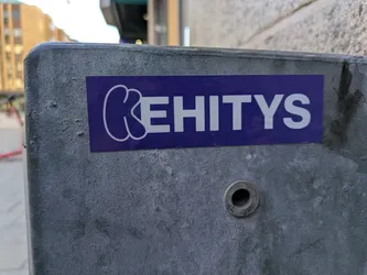 A rectangular sticker is affixed to a gray metal surface. The sticker is purple with white lettering that spells out 'KEHITYS.' The first letter 'K' is stylized with rounded, almost bubble-like shapes, while the remaining letters are in a simpler, sans-serif font.