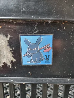 A square sticker featuring a dark-colored bunny rabbit wielding a bloody axe. The background is light blue. The artist's logo is visible in the bottom right corner: "Bunny Brigade". The sticker is affixed to a dirty, dark gray surface that appears to be metal or plastic.