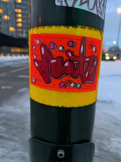 A close-up shot of a sticker on a pole. The sticker features red and white graffiti-style artwork on an orange background. There are also small white and pink dots scattered across the design. The sticker is surrounded by a yellow border and is attached to a dark green pole.