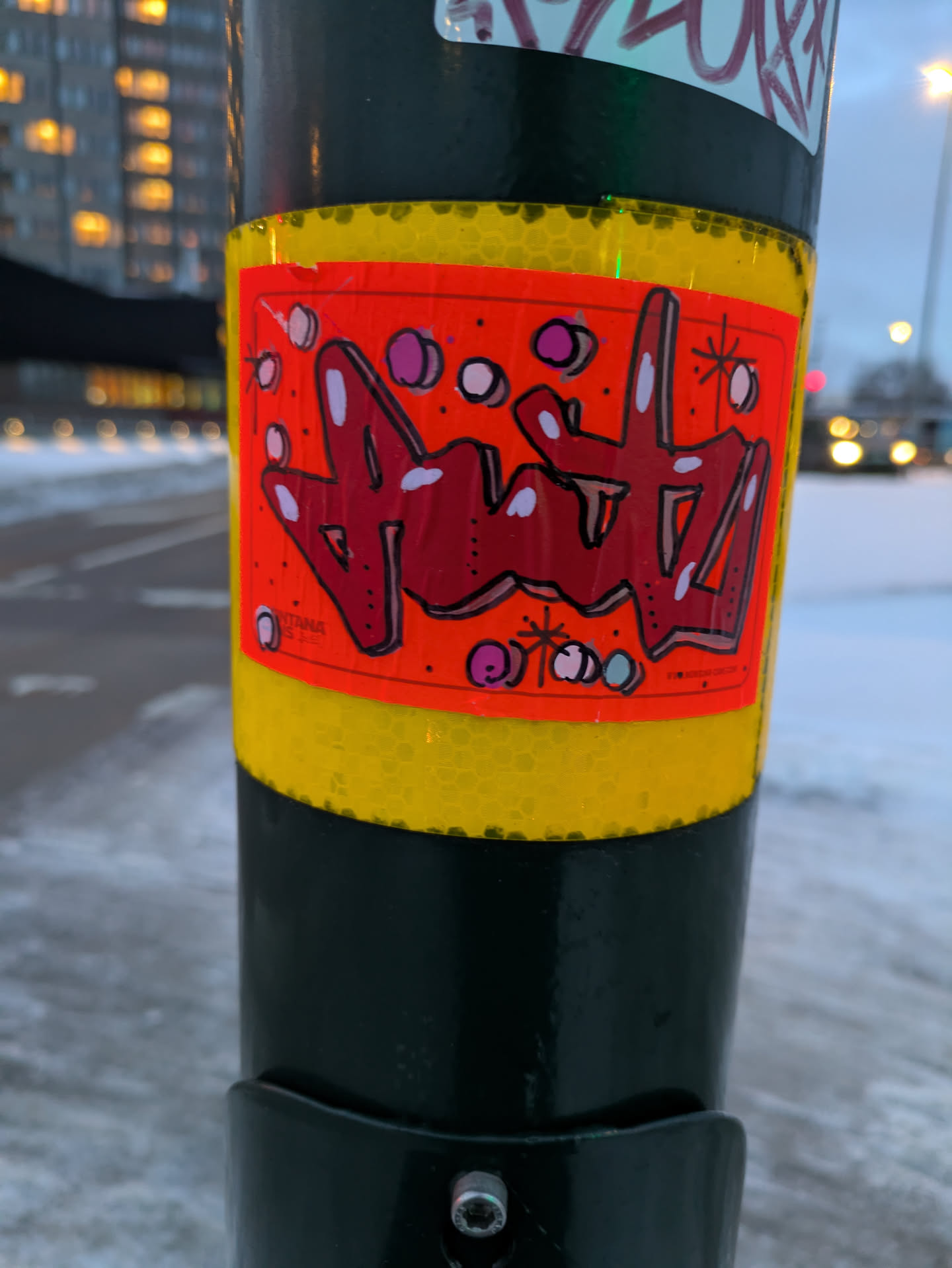 A close-up shot of a sticker on a pole. The sticker features red and white graffiti-style artwork on an orange background. There are also small white and pink dots scattered across the design. The sticker is surrounded by a yellow border and is attached to a dark green pole.