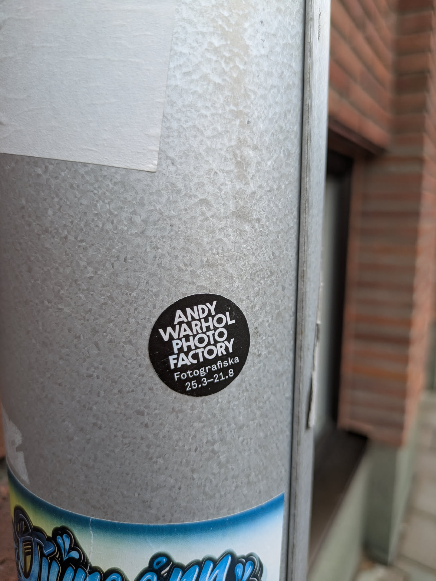 The sticker is circular with a black background and white text. The main text, 'ANDY WARHOL PHOTO FACTORY', is prominently displayed in a bold, sans-serif font, stacked over three lines. Below this, in a slightly smaller font, are the words 'Fotografiska' and the dates '25.3-21.8', indicating an exhibition period. The design is minimalist and clean, focusing solely on the text content. The sticker is affixed to a textured, galvanized metal pole.