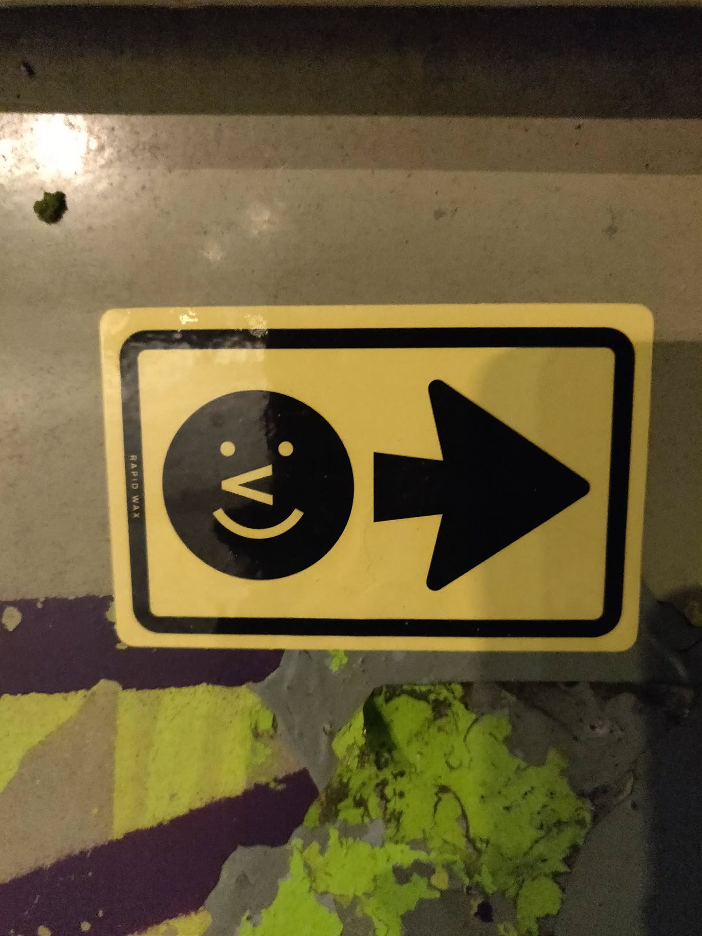 A yellow rectangular sticker with a black arrow pointing to the right and a simple smiling face to its left. The sticker has a slightly worn appearance, suggesting it may be placed outdoors. 