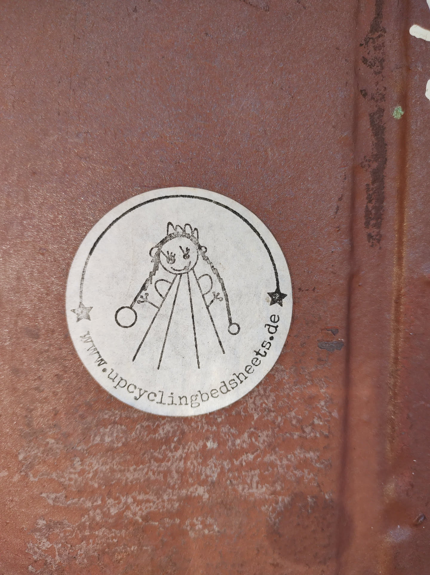 Round silver sticker with a drawing of a little girl and the website address www.upcyclingbedsheets.de. The sticker is on a rusty surface.