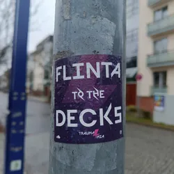 A sticker featuring the text "FLINTA to the DECKS" in white sans-serif font on a dark purple background with a lightning bolt and the word "TRAUMA MIA". The sticker is affixed to a metal pole outdoors and shows water droplets.
