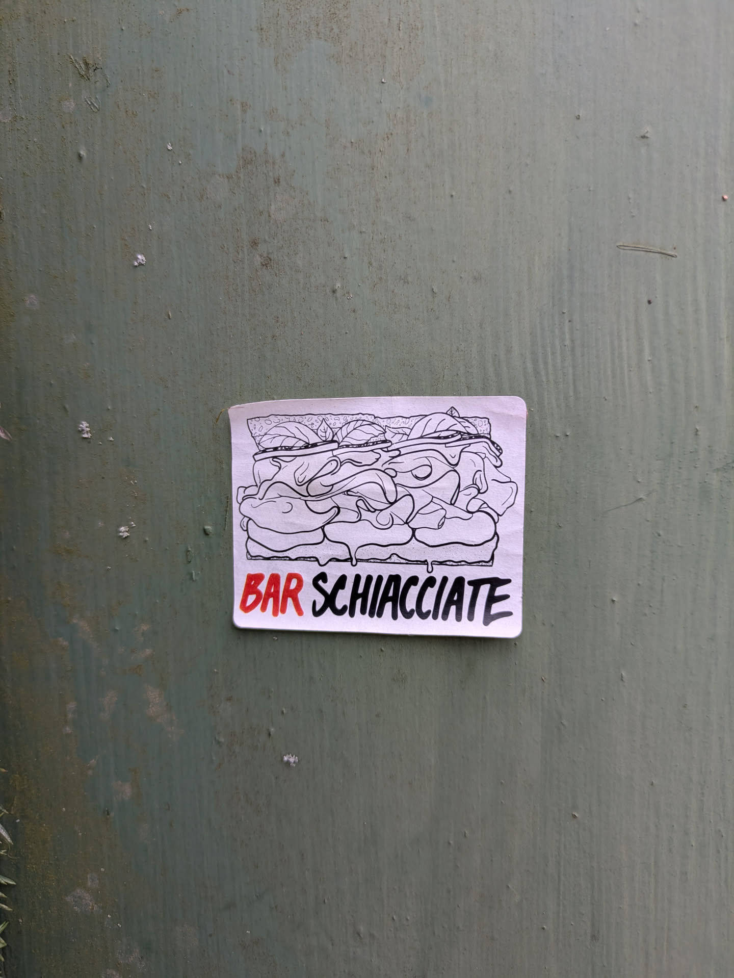 This sticker features a hand-drawn illustration of a schiacciata sandwich with ingredients spilling out. The words "BAR SCHIACCIATE" are printed in red underneath the sandwich. The sticker is adhered to a green background. The artwork has a homemade feel, hinting at a local business.