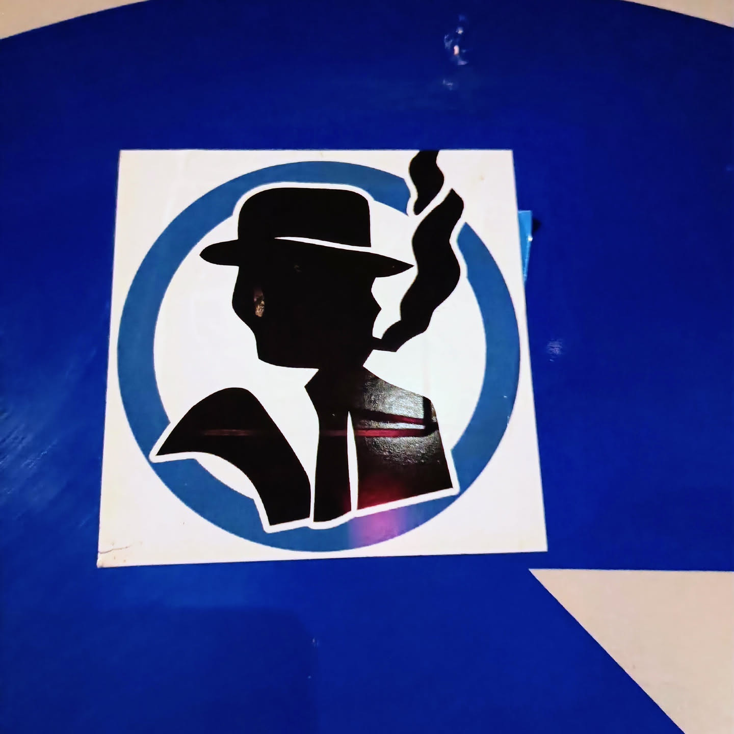 This sticker features a stylized silhouette of a man wearing a fedora and smoking. The design is simple yet striking, with a retro feel. The man is depicted in black against a white and light-blue circular background. The sticker is neatly placed on a blue surface.