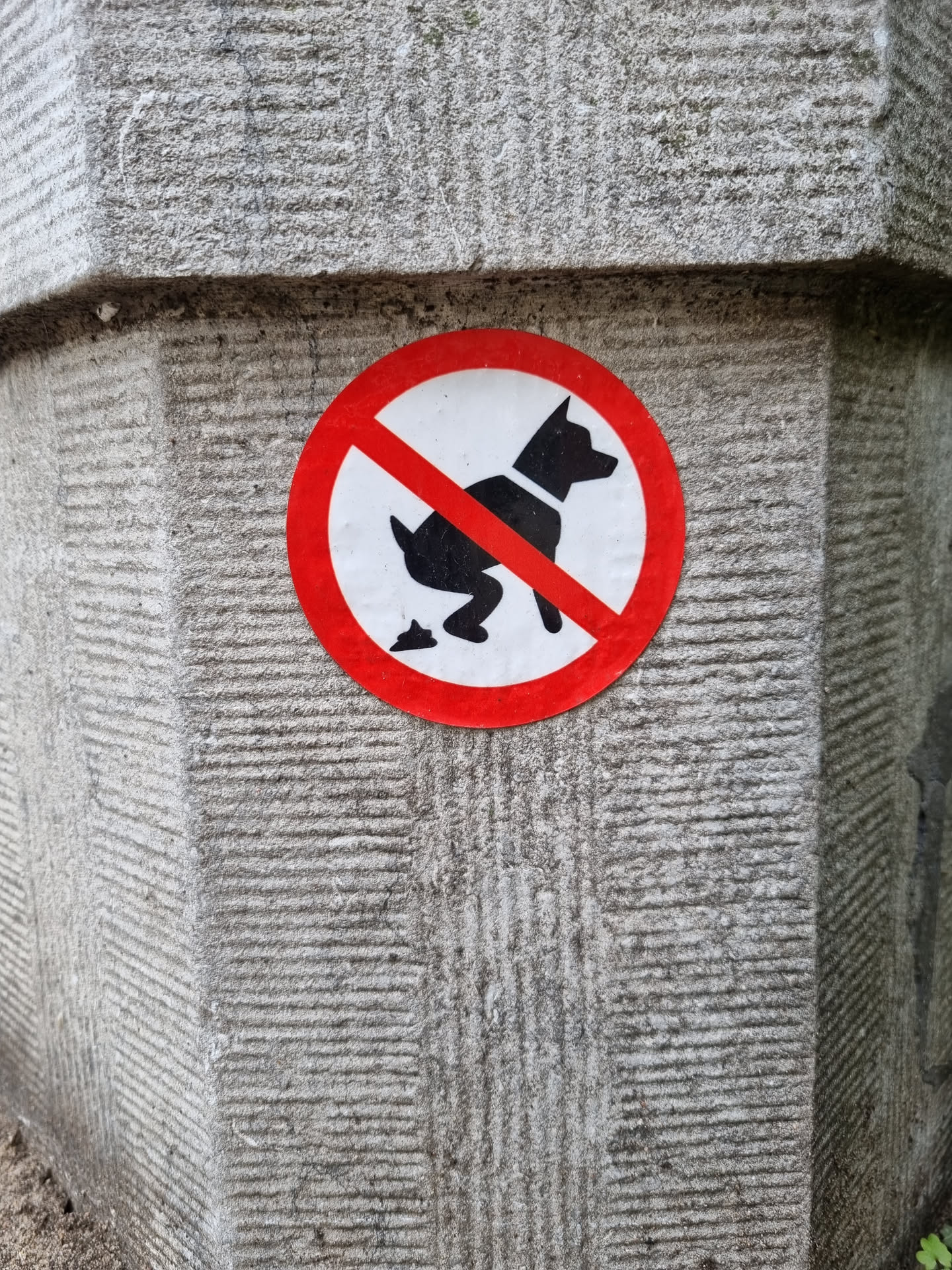 A round sticker with a red circle and a diagonal red line across a black silhouette of a dog pooping.  The sticker is affixed to a grey stone pillar.