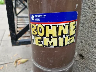 A yellow and black sticker with the words "BOHNE EMIL" written in a stylized font. There's a small heart symbol before the word "EMIL" and the letters "HK" in the bottom right corner. It appears to be affixed to a brown metal post.  A Priority Mail sticker is visible behind it.