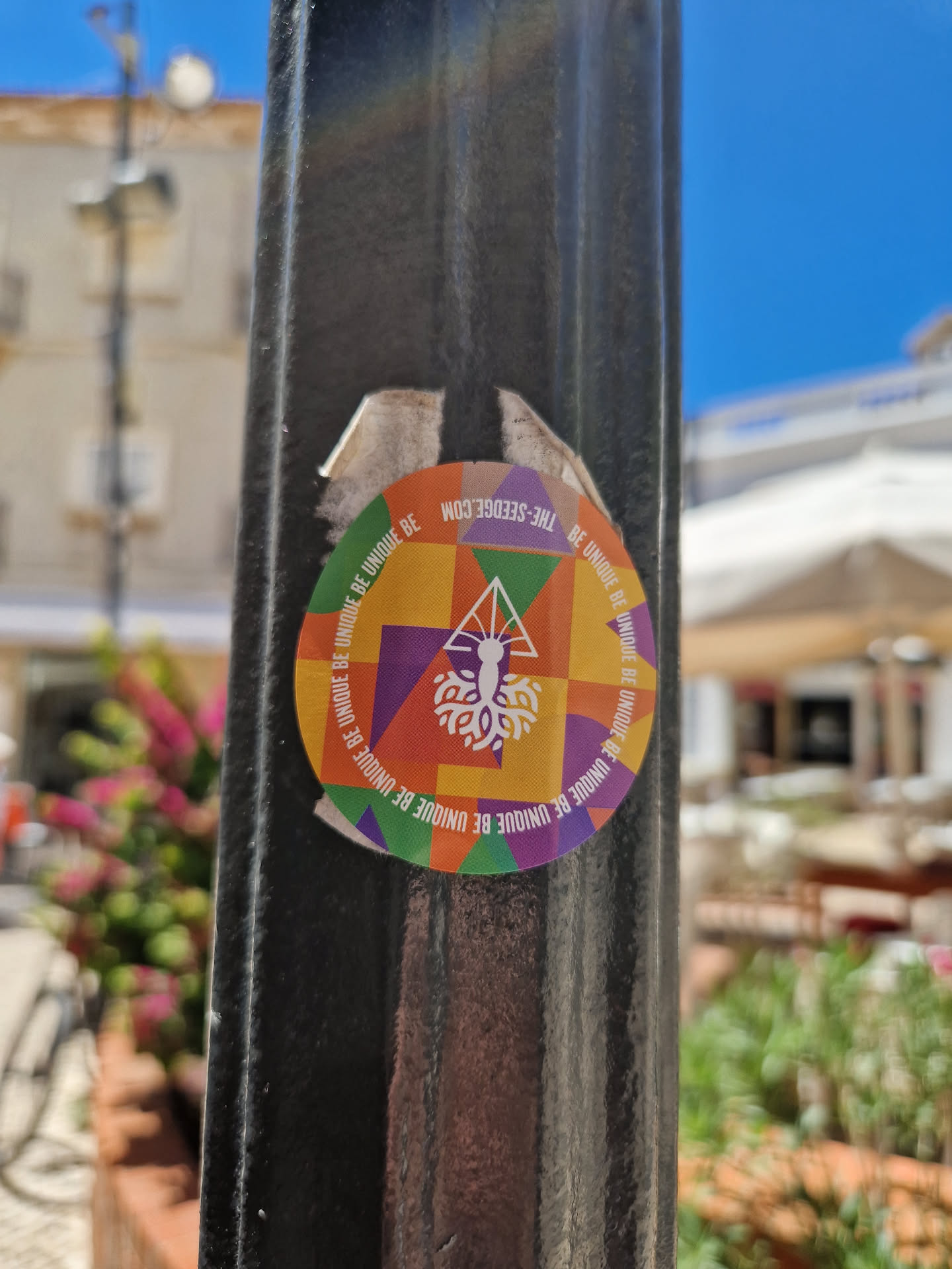 Round sticker with a geometric design in bright colors. The center features a tree-like image with text that repeats around the edge: "BE UNIQUE BE UNIQUE BE UNIQUE BE UNIQUE..."
