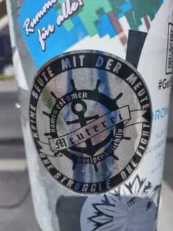 Silver circular sticker with a nautical theme. It features an anchor, a ship's wheel, and text in German that translates to "No loot with the mob. Name is omen. Kneipenhollektiv (Pub collective). One struggle, one fight."