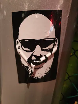 A black and white sticker featuring a stylized portrait of a bald man wearing sunglasses. The art style is reminiscent of stencil work or screen printing. The sticker is affixed to a metallic surface outdoors.