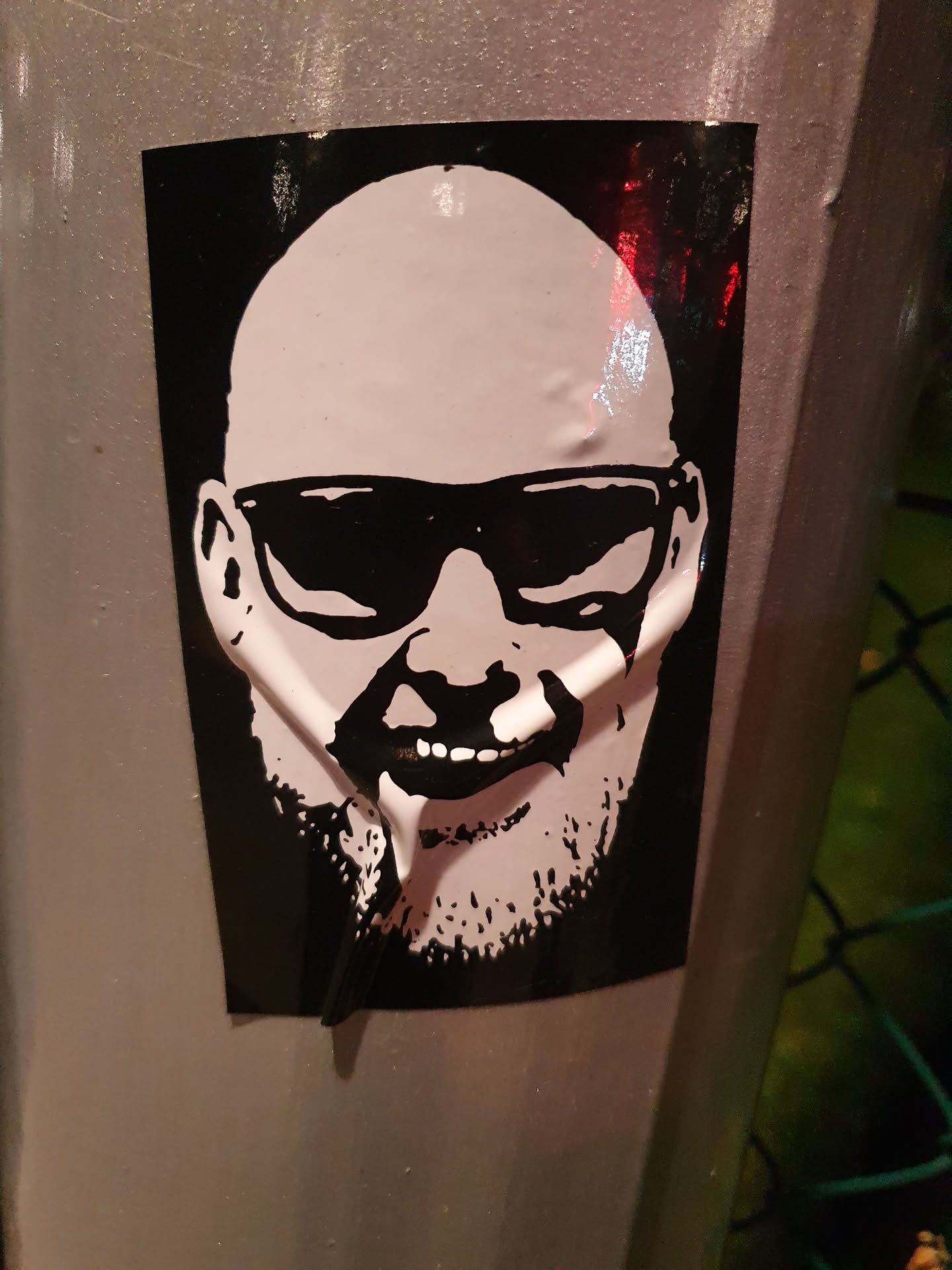 A black and white sticker featuring a stylized portrait of a bald man wearing sunglasses. The art style is reminiscent of stencil work or screen printing. The sticker is affixed to a metallic surface outdoors.