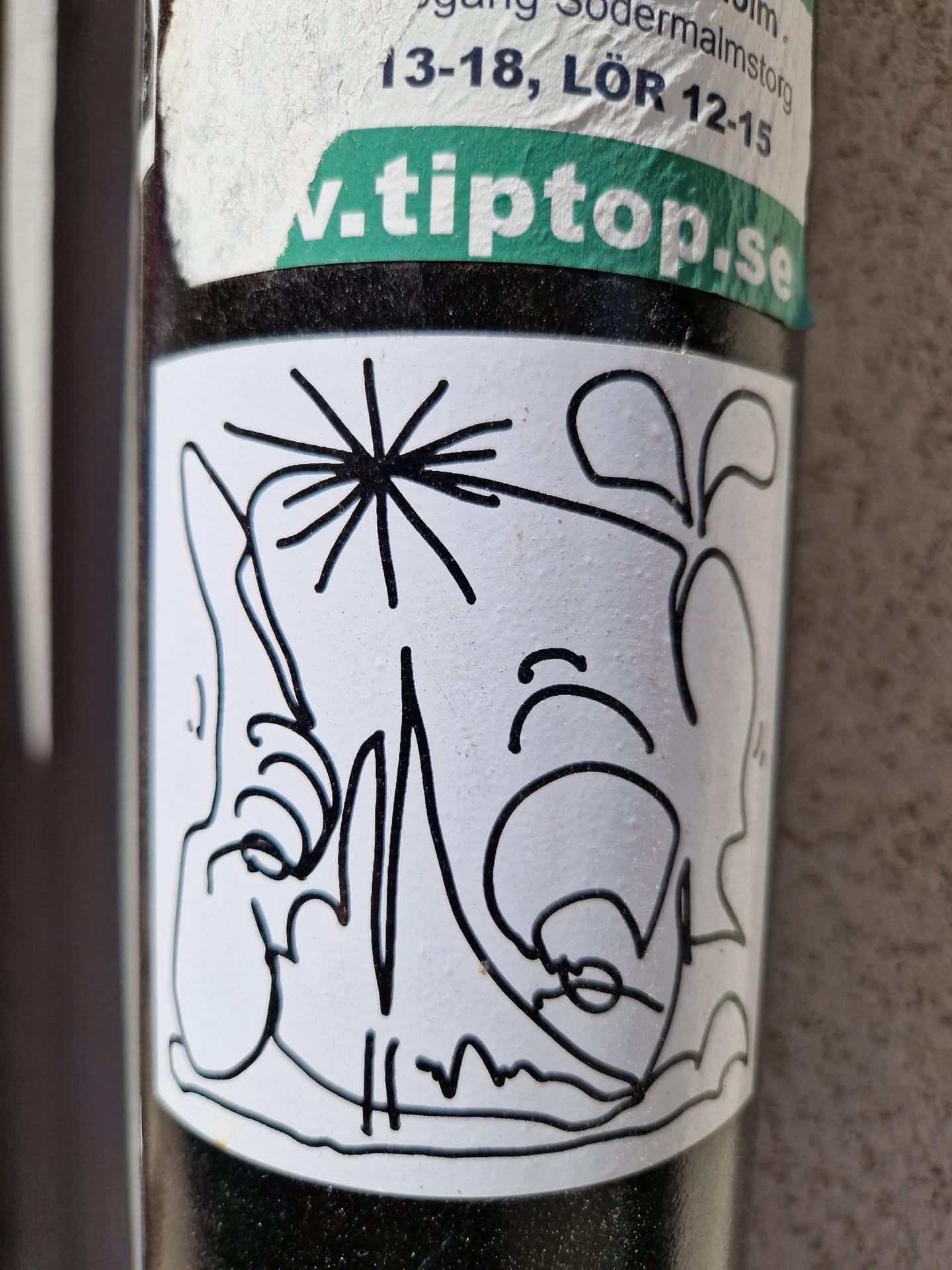 The sticker features a simple, elegant design. The main focus is a dandelion with its seeds dispersed, and next to it there is a smaller plant. The lines are clean and precise, creating a sense of simplicity and sophistication. The sticker is affixed to a dark surface and is slightly worn. 