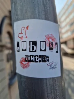 A white sticker with the word "МUВUКI vintage" written in a stylized font.  There are drawings of lips and a heart with an arrow. There's also a small signature at the bottom. 