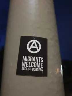 A black and white sticker with an anarchy symbol and the text "Migrants Welcome Abolish Borders".