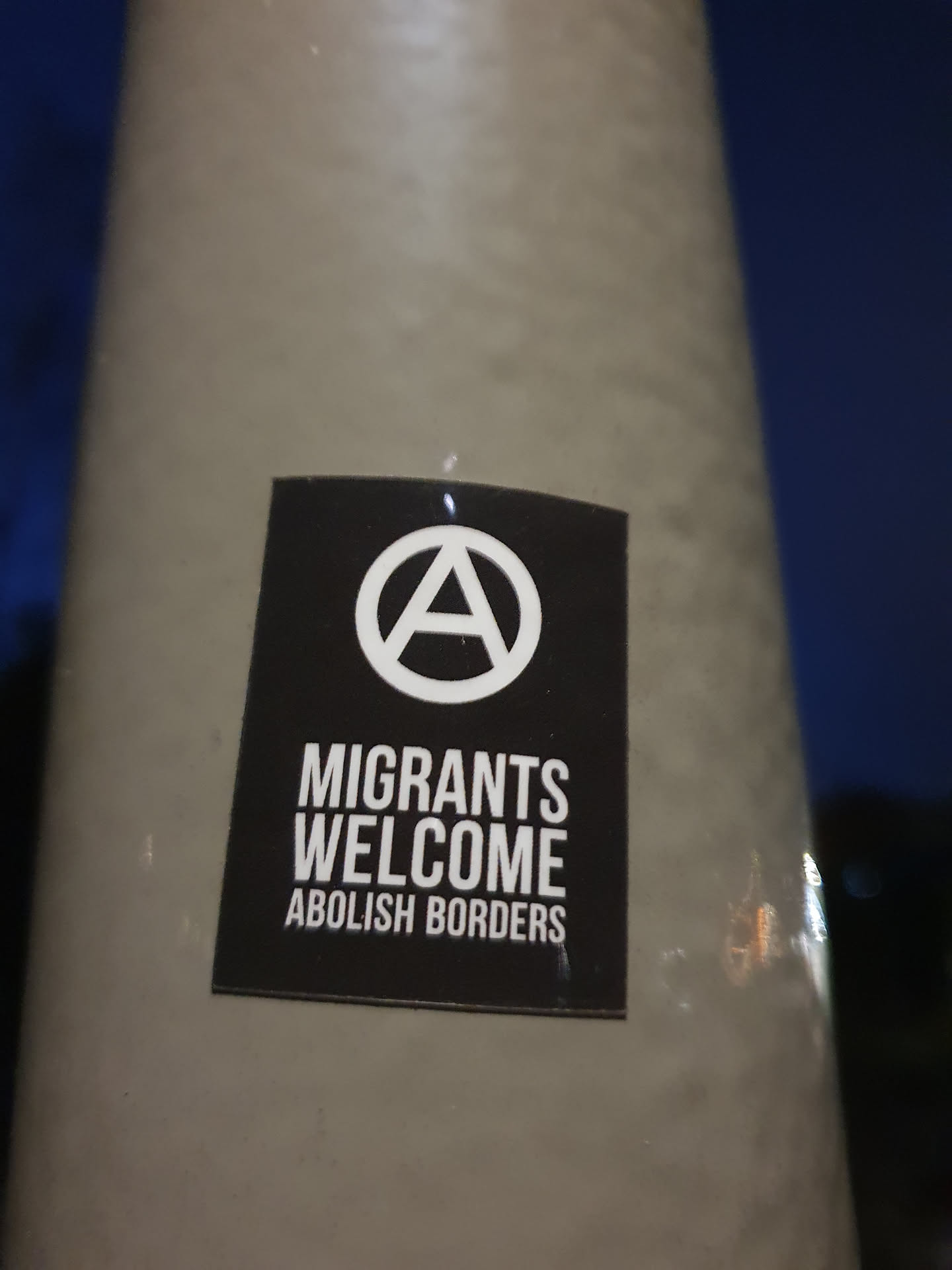 A black and white sticker with an anarchy symbol and the text "Migrants Welcome Abolish Borders".