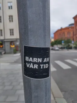 A black and white sticker on a metal pole. The text on the sticker says "Barn av vår tid" which translates to "Children of our time" in Swedish.