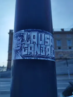 A sticker featuring a stylized illustration of a person smoking, with the text "LAUSANNE GANJA LIFE" prominently displayed. The sticker has a bluish-grey color scheme and appears to be affixed to a dark-colored pole outdoors.