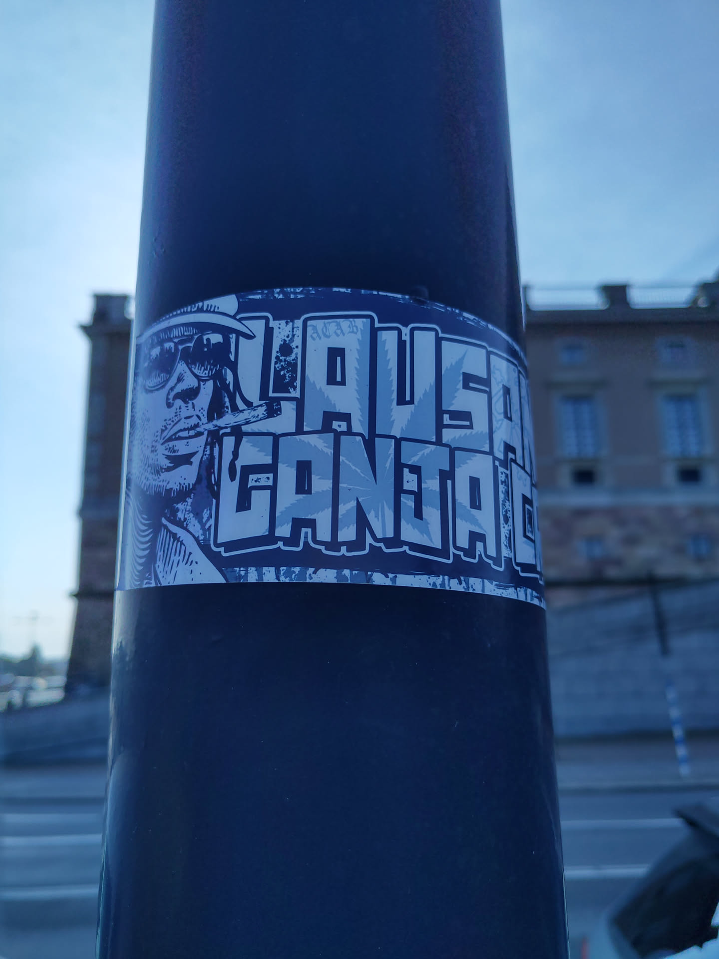 A sticker featuring a stylized illustration of a person smoking, with the text "LAUSANNE GANJA LIFE" prominently displayed. The sticker has a bluish-grey color scheme and appears to be affixed to a dark-colored pole outdoors.