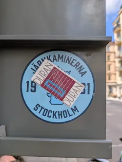 Street sticker The central image features two distinct stickers overlaid on a dark olive-green utility box or pillar. The primary, larger sticker is circular with a light blue background and a dark blue outer border. Dark blue text arcs around the top, "JÄRNKAMINERNA," and bottom, "STOCKHOLM." The numbers "19" and "31" flank a stylized blue graphic of what appears to be a stove or chimney in the center. Partially covering this, at a slight angle, is a smaller, rectangular sticker with a white border. This smaller sticker has a maroon interior featuring a grid of thin white lines, two thick light blue diagonal lines, and two thick light blue horizontal lines. The word "DURAN" is written vertically in an outline font on both the left and right sides of this maroon section.