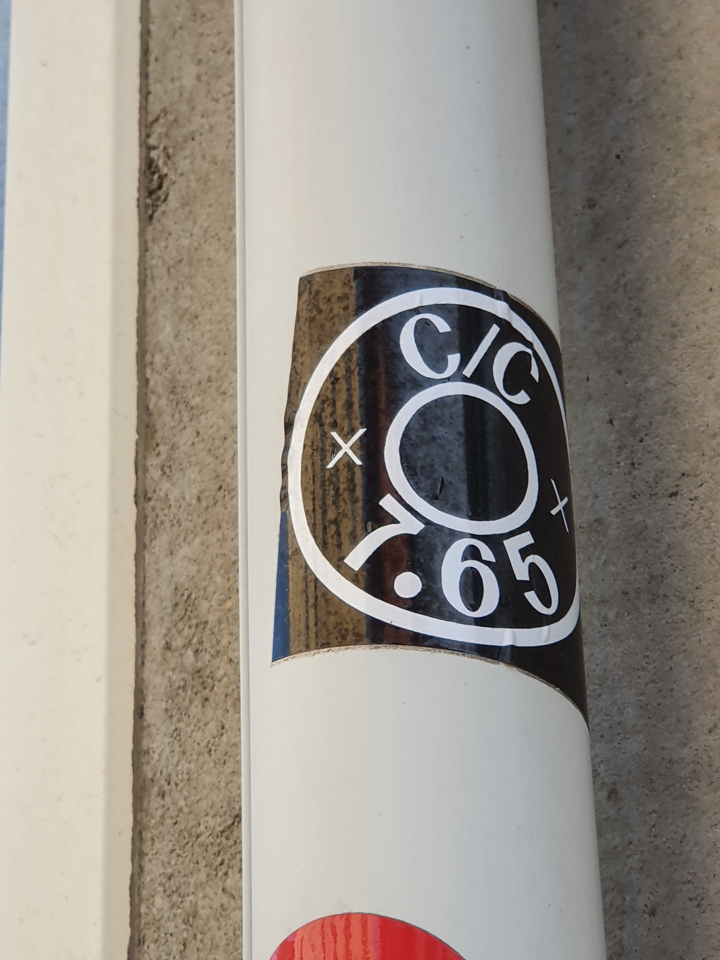 A black and white circular sticker with the text 'C/C' above a central circle and '7.65' below.  The sticker is affixed to a light beige cylindrical object.
