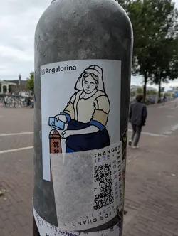 The sticker features a stylized, almost cartoonish illustration of a person dressed in historical attire. The figure has a serene expression and is depicted pouring a blue substance from a small rectangular container into a brown object adorned with three 'X' symbols. The illustration is outlined in black with light yellow and blue accents. Below the illustration, a QR code is prominent, surrounded by text. The sticker has been partially torn at the bottom.