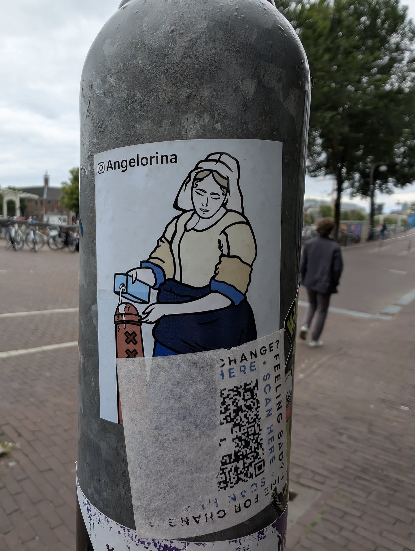 The sticker features a stylized, almost cartoonish illustration of a person dressed in historical attire. The figure has a serene expression and is depicted pouring a blue substance from a small rectangular container into a brown object adorned with three 'X' symbols. The illustration is outlined in black with light yellow and blue accents. Below the illustration, a QR code is prominent, surrounded by text. The sticker has been partially torn at the bottom.