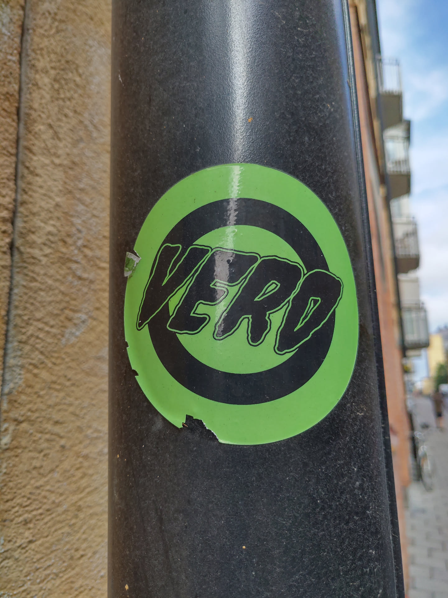A green and black sticker with the word VERO in a stylized font within a circle. The sticker is affixed to a dark gray pole, and shows some signs of wear and tear.