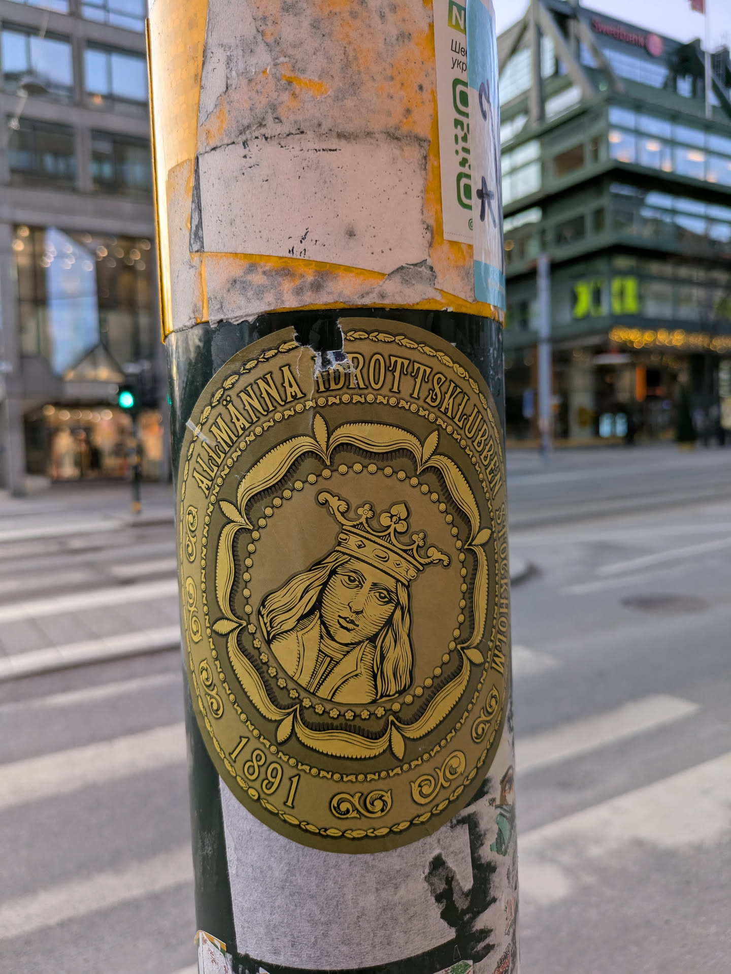 This round sticker features a gold design with an image of a crowned person, likely a historical figure,  and the text "Allmänna Idrottsklubben 1891". It appears to be affixed to a pole in an urban setting.
