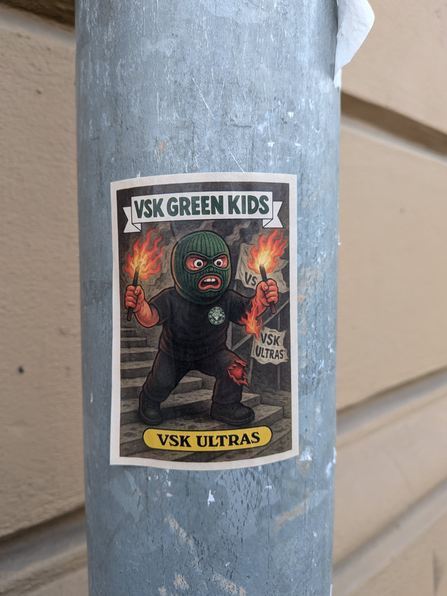 The sticker features a cartoonish character wearing a green balaclava and black clothing, with an angry expression. The character is holding a lit flare in its right hand, with flames erupting. The character's left hand is also depicted with flames. The background shows a set of stairs and two banners, one with 'VS' and another with 'VSK ULTRAS'. The overall color scheme is dark with vibrant orange and red flames. The sticker has a white border and a yellow oval at the bottom with text.
