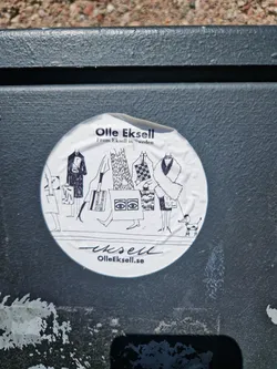 Round sticker featuring a black and white illustration of people shopping and a dog. The text includes the artist's name, origin and website.