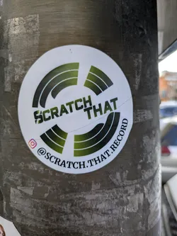 This is a white, round sticker with the words 'Scratch That' in bold black letters stacked on top of 'Record'. A green gradient symbol resembling rotating records surrounds the text. An Instagram icon is present with the username @scratch.that.record.